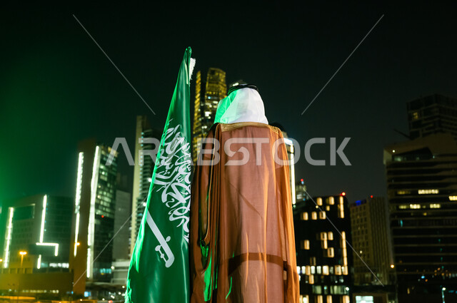 احياء ذكرى اليوم الوطني السعودي 23 سبتمبر،  الاحتفال بيوم العلم السعودي 11 مارس، الوقوف لتأمل الأبراج وناطحات السحاب المضاءة ليلا في مدينة الرياض، صورة من الخلف لرجل عربي خليجي سعودي يرتدي الغترة والبشت يقف بجانب العلم.