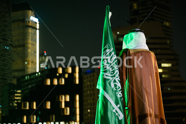 العيد الوطني 23 سبتمبر، الوقوف لتأمل الأبراج وناطحات السحاب المضاءة ليلا في مدينة الرياض، إحياء ذكرى اليوم الوطني السعودي، الاحتفال بيوم العلم السعودي 11 مارس، صورة من الخلف لرجل عربي خليجي سعودي يرتدي الغترة والبشت يقف بجانب العلم.