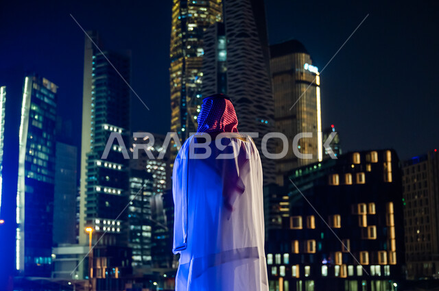 الاستمتاع والخروج في الهواء الطلق, الوقوف لتأمل التطور العمراني ومعالم مدينة الرياض, ارتداء الأزياء الشعبية التقليدية، صورة من الخلف لرجل عربي خليجي سعودي يرتدي الشماغ والبشت, المدينة المالية الاقتصادية المضاءة ليلا.