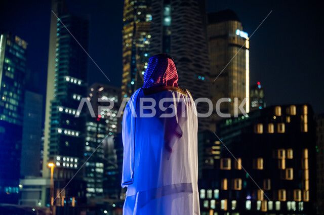 الاستمتاع والخروج في الهواء الطلق, الوقوف لتأمل التطور العمراني ومعالم مدينة الرياض, ارتداء الأزياء الشعبية التقليدية، صورة من الخلف لرجل عربي خليجي سعودي يرتدي الشماغ والبشت, المدينة المالية الاقتصادية المضاءة ليلا.