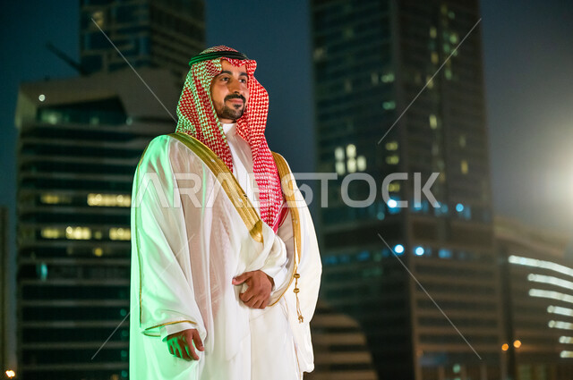 الأبراج وناطحات السحاب المضاءة ليلا في مدينة الرياض، ارتداء زي المناسبات الشعبية التقليدية، الفخر والاعتزاز بالانتماء للهوية السعودية, الوقوف بثقة والنظر إلى شيء ما, رجل عربي خليجي سعودي يرتدي الشماغ والبشت الأبيض.