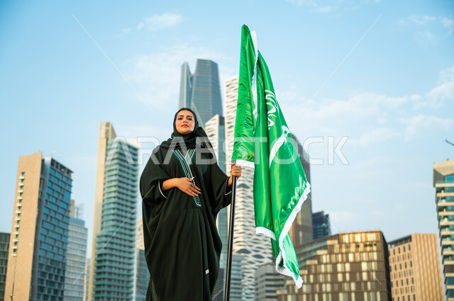 الوقوف بكبرياء وتجسيد مشاعر الولاء للدولة, الاحتفال بالعيد الوطني السعودي, اليوم الوطني في 8 ربيع الأول, إمرأة عربية سعودية خليجية محجبة ترتدي العباءة وتمسك علم المملكة بفخر وتقدير, يوم العلم السعودي 11 مارس, خلفية أبراج كافد في الرياض