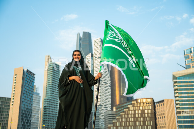 مشاعر الحب والولاء للسعودية, يوم الوحدة والتاريخ الحديث للمملكة, الاحتفال باليوم الوطني السعودي 8 ربيع الأول, إمرأة عربية سعودية خليجية محجبة مبتسمة ترتدي العباءة وتمسك علم المملكة بفخر وتقدير, يوم العلم السعودي 11 مارس, خلفية أبراج كافد في الرياض
