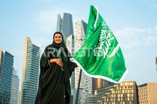 يوم الوحدة والتاريخ الحديث للمملكة, تجسيد مشاعر الحب والولاء, الاحتفال باليوم الوطني السعودي  8 ربيع الأول, إمرأة عربية سعودية خليجية محجبة ترتدي العباءة وتمسك علم المملكة بفخر وتقدير, يوم العلم السعودي 11 مارس, خلفية أبراج كافد في الرياض