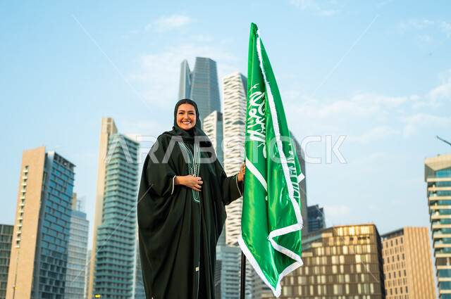 Embodying feelings of love and loyalty, Day of Unity and the modern history of the Kingdom, Celebrating Saudi National Day September 23, A veiled Saudi Gulf Arab woman wearing the abaya and holding the Kingdom’s flag with pride and appreciation, Saudi Flag Day March 11, Background of the Kafed Towers in Riyadh