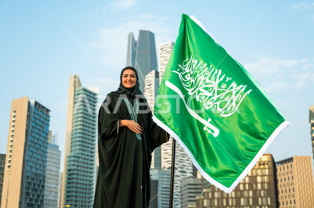 الاحتفال باليوم الوطني السعودي 23 سبتمبر, تجسيد مشاعر الحب والولاء, يوم الوحدة والتاريخ الحديث للمملكة, إمرأة عربية سعودية خليجية محجبة ترتدي العباءة وتمسك علم المملكة بفخر وتقدير, يوم العلم السعودي 11 مارس, خلفية أبراج كافد في الرياض