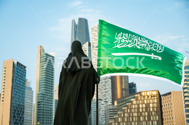 الحب والولاء للسعودية بالمشاركة باليوم الوطني السعودي 23 سبتمبر, صورة من الخلف لإمرأة عربية سعودية خليجية محجبة ترتدي العباءة تمسك علم المملكة بفخر وتقدير, يوم العلم السعودي 11 مارس, خلفية أبراج كافد في الرياض