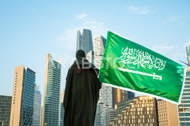 الولاء للسعودية بالمشاركة باليوم الوطني السعودي 23 سبتمبر, صورة من الخلف لإمرأة عربية سعودية خليجية محجبة ترتدي العباءة تمسك علم المملكة بفخر وتقدير, يوم العلم السعودي 11 مارس, خلفية أبراج كافد في الرياض