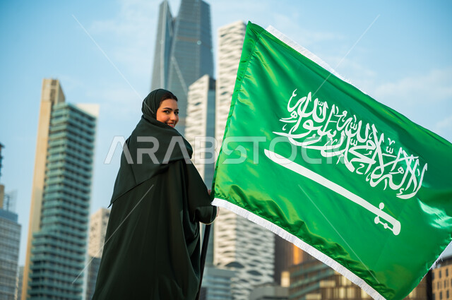 النظر إلى الكاميرا بإيماءات السعادة, الانتماء للوطن والمشاركة بالمناسبات الوطنية, إمرأة عربية سعودية خليجية مبتسمة ترتدي العباءة والحجاب تمسك علم المملكة باحترام وتقدير, استذكار العيد الوطني, ذكرى اليوم الوطني في 23 سبتمبر, يوم العلم السعودي 11 مارس