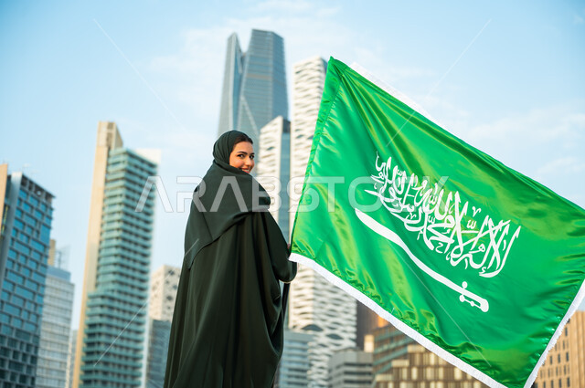 الانتماء للوطن والمشاركة بالمناسبات الوطنية, إمرأة عربية سعودية خليجية مبتسمة ترتدي العباءة والحجاب تمسك علم المملكة باحترام وتقدير, استذكار العيد الوطني, ذكرى اليوم الوطني في 23 سبتمبر, يوم العلم السعودي 11 مارس, النظر إلى الكاميرا بإيماءات السعادة