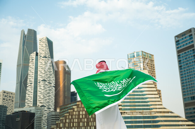 الحفاظ على الهوية الوطنية السعودية, الاحتفال بذكرى اليوم الوطني 23 سبتمبر, استذكار يوم العلم السعودي 11 مارس, صورة من الخلف لرجل عربي سعودي خليجي يرتدي العلم بحب وولاء للمملكة, خلفية الأبراج المالية والمباني الهندسية الحديثة