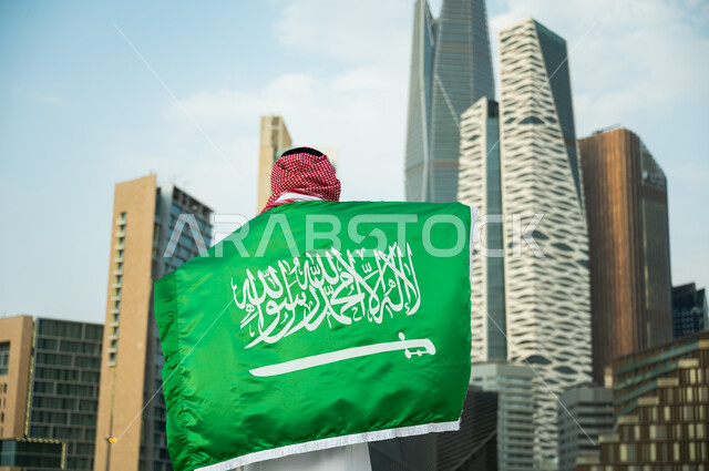 استذكار يوم العلم السعودي 11 مارس, الحفاظ على الهوية الوطنية السعودية, الاحتفال بذكرى اليوم الوطني  23 سبتمبر, صورة من الخلف لرجل عربي سعودي خليجي يرتدي العلم بحب وولاء للمملكة, خلفية الأبراج المالية والمباني الهندسية الحديثة