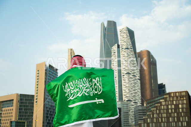 الحفاظ على الهوية الوطنية السعودية, الاحتفال بذكرى اليوم الوطني 23 سبتمبر, استذكار يوم العلم السعودي 11 مارس, صورة من الخلف لرجل عربي سعودي خليجي يرتدي العلم بحب وولاء للمملكة, خلفية الأبراج المالية والمباني الهندسية الحديثة