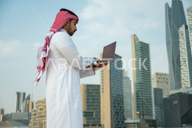 التطبيقات الالكترونية الحديثة وفرص العمل على الانترنت, عقد صفقات عمل ناجحة مع رجال أعمال عبر الأونلاين, العمل عن بعد عبر مواقع التواصل الاجتماعي, رجل عربي سعودي خليجي يرتدي الثوب والشماغ الأحمر يقوم باستخدام جهاز الحاسوب, خلفية أبراج الملك عبدالله المالية