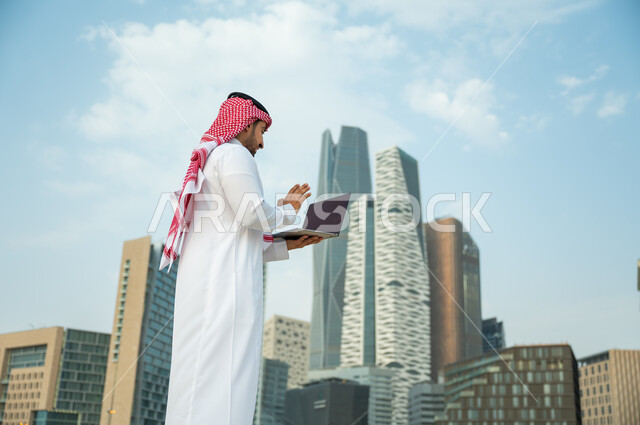 صداقات وحوارات عبر مواقع التواصل الاجتماعي, التطور العمراني في مدينة الرياض, الدردشة والتواصل مع الأصدقاء وإجراء مكالمة فيديو, رجل عربي سعودي خليجي يرتدي الثوب السعودي والشماغ يلوح بيده لإلقاء التحية عبر جهاز الحاسوب, خلفية أبراج كافد المالية وضح النهار