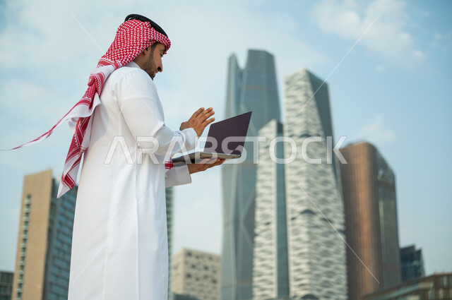 الدردشة والتواصل مع الأصدقاء وإجراء مكالمة فيديو, التطور العمراني في مدينة الرياض, رجل عربي سعودي خليجي يرتدي الثوب السعودي والشماغ يلوح بيده لإلقاء التحية عبر جهاز الحاسوب, صداقات وحوارات عبر مواقع التواصل الاجتماعي, خلفية أبراج كافد المالية وضح النهار