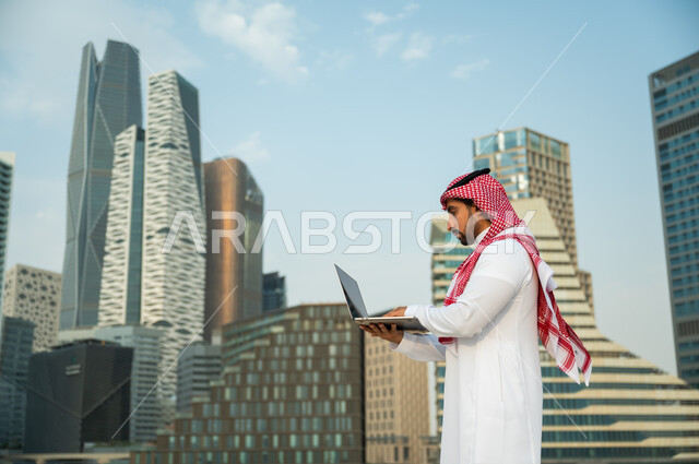 العمل عن بعد عبر مواقع التواصل الاجتماعي, عقد صفقات عمل ناجحة مع رجال أعمال عبر الأونلاين, رجل عربي سعودي خليجي يرتدي الثوب والشماغ الأحمر يقوم باستخدام جهاز الحاسوب, التطبيقات الالكترونية الحديثة وفرص العمل على الانترنت, خلفية أبراج الملك عبدالله المالية