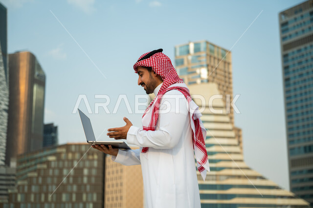 العمل عن بعد عبر مواقع التواصل الاجتماعي, عقد صفقات عمل ناجحة مع رجال أعمال عبر الأونلاين, رجل عربي سعودي خليجي يرتدي الثوب والشماغ الأحمر يقوم بإجراء مكالمة فيديو عبر الحاسوب, استخدام التطبيقات الالكترونية الحديثة, خلفية أبراج الملك عبدالله المالية