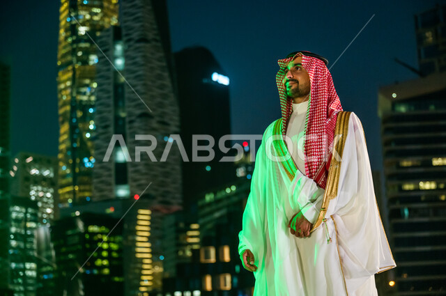 ارتداء أزياء المناسبات والاحتفالات الشعبية التقليدية، التباهي والفخر بالهوية السعودية, خلفية الأبراج المالية الاقتصادية المضاءة ليلا في مدينة الرياض، الوقوف بثقة والنظر إلى شيء ما, رجل عربي خليجي سعودي مبتسم يرتدي الشماغ والبشت الأبيض.