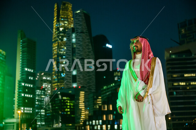 ارتداء أزياء المناسبات والاحتفالات الشعبية التقليدية، التباهي والفخر بالهوية السعودية, خلفية الأبراج المالية الاقتصادية المضاءة ليلا في مدينة الرياض، الوقوف بثقة والنظر إلى شيء ما, رجل عربي خليجي سعودي مبتسم يرتدي الشماغ والبشت الأبيض.