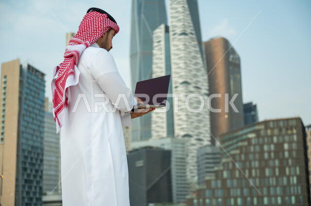 استخدام الأجهزة الالكترونية الحديثة, عقد صفقات عمل ناجحة مع رجال أعمال عبر الأونلاين, العمل عن بعد عبر مواقع التواصل الاجتماعي, صورة مقربة لرجل عربي سعودي خليجي يرتدي الثوب والشماغ يقوم يقوم بالبحث عن معلومة ما على الانترنت, أبراج الملك عبدالله المالية