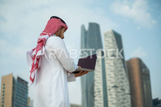 Searching for information on the Internet, working remotely via social media, a Saudi Arabian Gulf man wearing a red thobe and head scarf using a computer, modern electronic applications and job opportunities on the Internet, the background of King Abdull