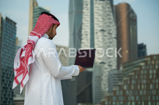 استخدام الأجهزة الالكترونية الحديثة, عقد صفقات عمل ناجحة مع رجال أعمال عبر الأونلاين, العمل عن بعد عبر مواقع التواصل الاجتماعي, صورة مقربة لرجل عربي سعودي خليجي يرتدي الثوب والشماغ يقوم يقوم بالبحث عن معلومة ما على الانترنت, أبراج الملك عبدالله المالية