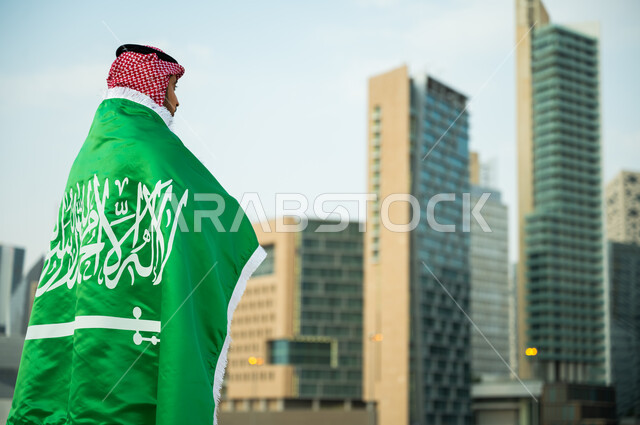 Standing proudly on National Day, September 23, wearing the Saudi flag with appreciation and respect, expressing love and loyalty on Flag Day, March 11, a Saudi Gulf Arab man wearing the traditional thobe and shemagh participating in Saudi national holidays and events