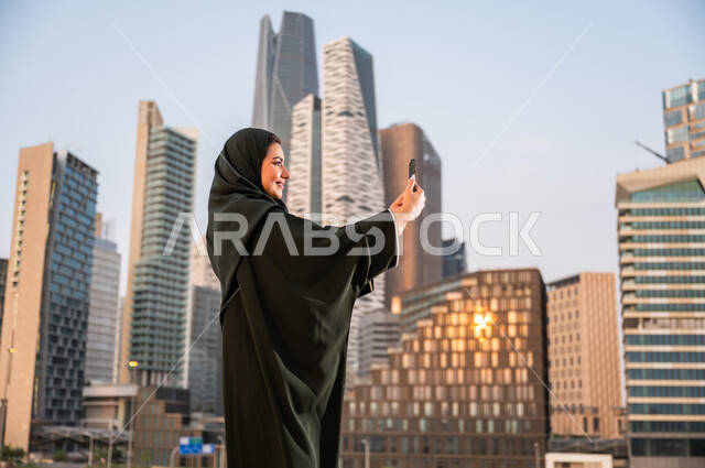 الاستمتاع في التقاط صور سيلفي تذكارية عبر الجوال, الخروج في الهواء الطلق في وقت النهار, سيدة عربية سعودية خليجية ترتدي العباءة والحجاب تقوم باستخدام الهاتف المحمول, خلفية أبراج الملك عبدالله المالية, فعاليات وأنشطة ترفيهية وقضاء وقت ممتع في مدينة الرياض