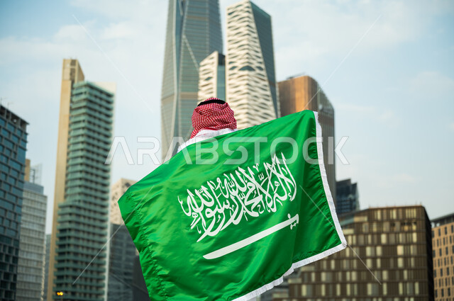 رفع العلم على الأكتاف احتفالا بالعيد الوطني , تمجيد اليوم الوطني في 23 سبتمبر, يوم العلم السعودي 11 مارس, صورة مقربة لرجل عربي سعودي خليجي يرتدي الزي التقليدي يشارك بالمناسبات والأعياد الوطنية, خلفية أبراج الملك عبدالله