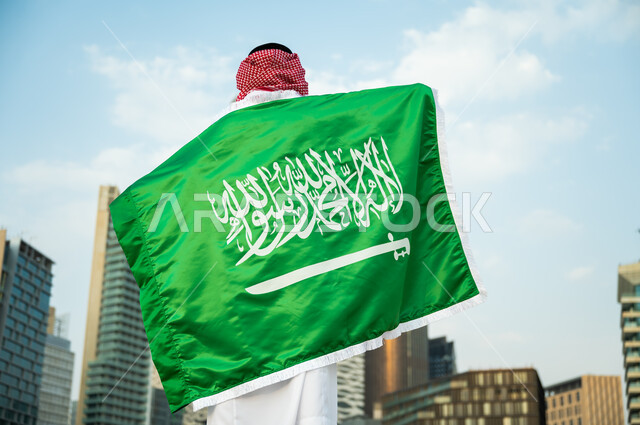 تمجيد اليوم الوطني في 23 سبتمبر, رفع العلم على الأكتاف احتفالا بالعيد الوطني, يوم العلم السعودي 11 مارس, صورة مقربة لرجل عربي سعودي خليجي يرتدي الزي التقليدي يشارك بالمناسبات والأعياد الوطنية, خلفية أبراج الملك عبدالله