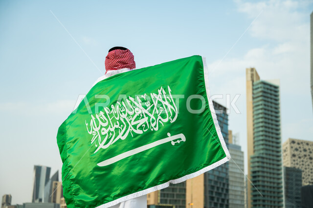 الاعتزاز والفخر بالهوية الوطنية السعودية, الاحتفال بذكرى اليوم الوطني 23 سبتمبر, استذكار يوم العلم السعودي 11 مارس, صورة من الخلف لرجل عربي سعودي خليجي يرتدي العلم بحب وولاء للمملكة, خلفية الأبراج المالية والمباني الهندسية الحديثة