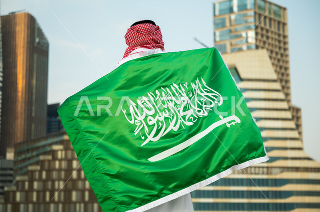 الفخر والاعتزاز بالهوية الوطنية السعودية, الاحتفال بذكرى اليوم الوطني 23 سبتمبر, استذكار يوم العلم السعودي 11 مارس, صورة من الخلف لرجل عربي سعودي خليجي يرتدي العلم بحب وولاء للمملكة, خلفية الأبراج المالية والمباني الهندسية الحديثة