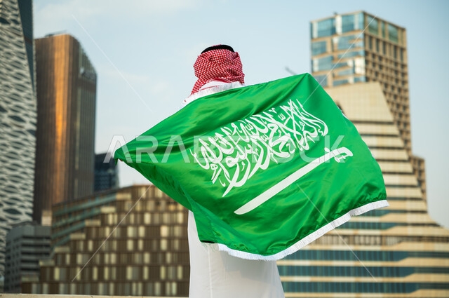 الشعور بالانتماء والحفاظ على الهوية الوطنية السعودية, الاحتفال بذكرى اليوم الوطني 23 سبتمبر, استذكار يوم العلم السعودي 11 مارس, صورة من الخلف لرجل عربي سعودي خليجي يرتدي العلم بحب وولاء للمملكة, خلفية الأبراج المالية والمباني الهندسية الحديثة