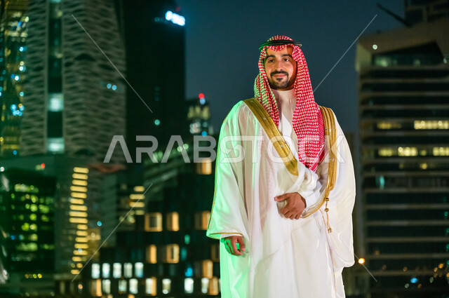 الاعتزاز بالدولة العربية السعودية، ارتداء أزياء المناسبات والاحتفالات الشعبية التقليدية، خلفية مركز الملك عبد الله المالي المضاءة ليلا في مدينة الرياض، الوقوف بثقة والنظر إلى شيء ما, رجل عربي خليجي سعودي يرتدي الشماغ والبشت الأبيض.