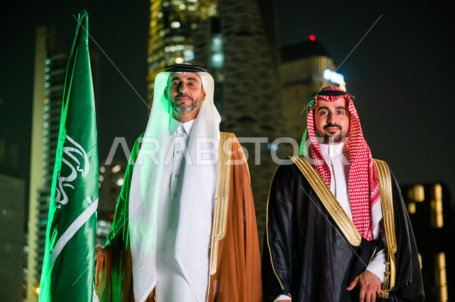 الاحتفال بذكرى اليوم الوطني  23 سبتمبر , أزياء المناسبات والاحتفالات الشعبية, التعبير عن الحب والولاء في يوم العلم 11 مارس, صورة مقربة لرجلين عربيين سعوديبن خليجيين يرتديان الزي السعودي التقليدي، الوقوف باعتزاز بالوطن، خلفية الأبراج والمباني في الرياض.