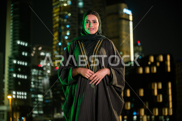 الاعتزاز والتباهي بالهوية السعودية، سيدة عربية خليجية سعودية محجبة ترتدي العباءة، خلفية الأبراج وناطحات السحاب المضاءة ليلا في مدينة الرياض، الوقوف بثقة والنظر إلى الكاميرا، إيماءات وجه تدل على السعادة. 