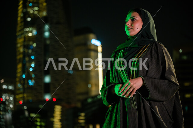 الاعتزاز والتباهي بالهوية السعودية، سيدة عربية خليجية سعودية محجبة ترتدي عباءة تقف أمام الأبراج وناطحات السحاب المضاءة ليلا في مدينة الرياض، الوقوف بثقة والنظر إلى شيء ما، الاهتمام بالمظهر الخارجي. 