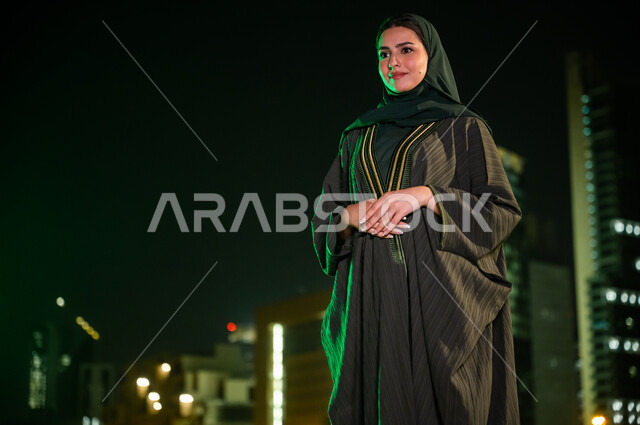 الاعتزاز والتباهي بالهوية السعودية، سيدة عربية خليجية سعودية محجبة ترتدي عباءة تقف أمام الأبراج وناطحات السحاب المضاءة ليلا في مدينة الرياض، الوقوف بثقة والنظر إلى شيء ما، إيماءات وجه تدل على السعادة. 