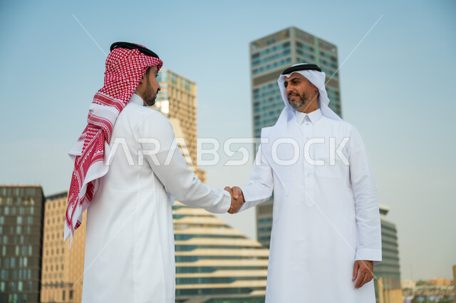 إيماءات تدل على السعادة بنجاح الصفقة, اتفاقات عمل بين الشركات الإعمارية, التعارف وبناء علاقات اجتماعية, إجراء صفقات عمل ناجحة, صورة مقربة لرجلين عربيين سعوديين خليجيين يرتديان الزي التقليدي يقومان بمصافحة بعضهما, خلفية أبراج مركز عبد الله المالي