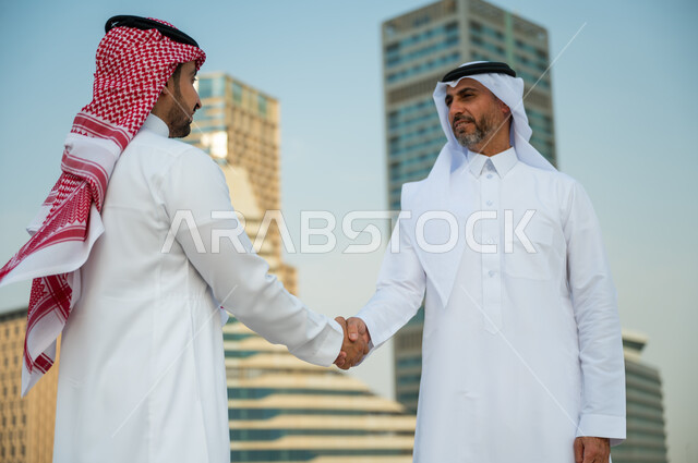 إيماءات تدل على السعادة بنجاح الصفقة, اتفاقات عمل بين الشركات الإعمارية, التعارف وبناء علاقات اجتماعية, إجراء صفقات عمل ناجحة, صورة مقربة لرجلين عربيين سعوديين خليجيين يرتديان الزي التقليدي يقومان بمصافحة بعضهما, خلفية أبراج مركز عبد الله المالي