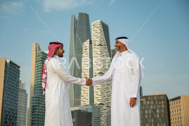 اتفاقات عمل بين الشركات الإعمارية, إيماءات تدل على السعادة بنجاح الصفقة, التعارف وبناء علاقات اجتماعية, إجراء صفقات عمل ناجحة بين رجال الأعمال, رجلان عربيان سعوديان خليجيان يرتديان الزي التقليدي يقومان بمصافحة بعضهما, خلفية أبراج مركز عبد الله المالي