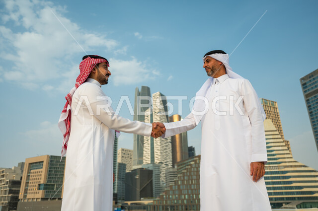 اتفاقات عمل بين الشركات الإعمارية, إيماءات تدل على السعادة بنجاح الصفقة, التعارف وبناء علاقات اجتماعية, إجراء صفقات عمل ناجحة بين رجال الأعمال, رجلان عربيان سعوديان خليجيان يرتديان الزي التقليدي يقومان بمصافحة بعضهما, خلفية أبراج مركز عبد الله المالي