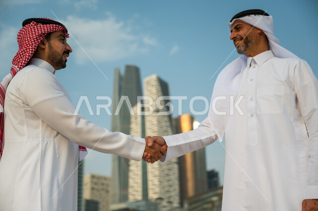 البهجة و السعادة بنجاح الصفقة, اتفاقات عمل بين الشركات الإعمارية, التعارف وبناء علاقات اجتماعية, إجراء صفقات عمل ناجحة, صورة مقربة لرجلين عربيين سعوديين خليجيين يرتديان الزي التقليدي يقومان بمصافحة بعضهما, خلفية أبراج مركز عبد الله المالي