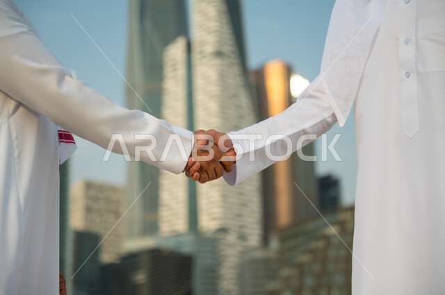 اتفاقات عمل بين الشركات الإعمارية, التعارف وبناء علاقات اجتماعية, إجراء صفقات عمل ناجحة بين رجال الأعمال, صورة مقربة ليد رجلين عربيين سعوديين خليجيين يرتديان الزي التقليدي يقومان بمصافحة بعضهما, خلفية أبراج مركز عبد الله المالي