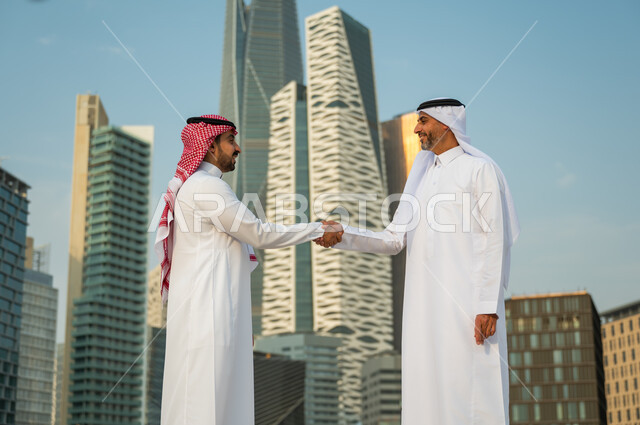 التعارف وبناء علاقات اجتماعية, إيماءات تدل على السعادة بنجاح الصفقة, اتفاقات عمل بين الشركات الإعمارية, إجراء صفقات عمل ناجحة بين رجال الأعمال, رجلان عربيان سعوديان خليجيان يرتديان الزي التقليدي يقومان بمصافحة بعضهما, خلفية أبراج مركز عبد الله المالي