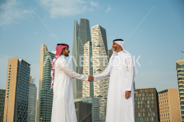 التعارف وبناء علاقات اجتماعية, إيماءات تدل على السعادة بنجاح الصفقة, اتفاقات عمل بين الشركات الإعمارية, إجراء صفقات عمل ناجحة بين رجال الأعمال, رجلان عربيان سعوديان خليجيان يرتديان الزي التقليدي يقومان بمصافحة بعضهما, خلفية أبراج مركز عبد الله المالي