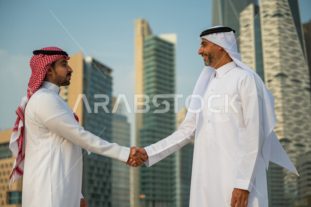 إجراء صفقات عمل ناجحة, إيماءات تدل على السعادة بنجاح الصفقة, اتفاقات عمل بين الشركات الإعمارية, التعارف وبناء علاقات اجتماعية, صورة مقربة لرجلين عربيين سعوديين خليجيين يرتديان الزي التقليدي يقومان بمصافحة بعضهما, خلفية أبراج مركز عبد الله المالي