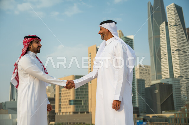 تكوين معارف وتوطيد العلاقات الاجتماعية, إيماءات تدل على السعادة بنجاح الصفقة, اتفاقات عمل بين الشركات الإعمارية, إجراء صفقات عمل بين رجال الأعمال, رجلان عربيان سعوديان خليجيان يرتديان الزي التقليدي يقومان بمصافحة بعضهما, خلفية أبراج مركز عبد الله المالي