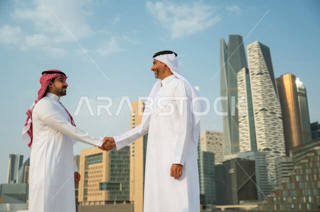 تكوين معارف وتوطيد العلاقات الاجتماعية, إيماءات تدل على السعادة بنجاح الصفقة, اتفاقات عمل بين الشركات الإعمارية, إجراء صفقات عمل بين رجال الأعمال, رجلان عربيان سعوديان خليجيان يرتديان الزي التقليدي يقومان بمصافحة بعضهما, خلفية أبراج مركز عبد الله المالي